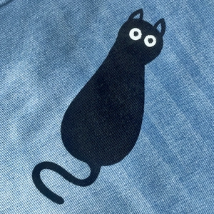 Détail de la broderie chat noir minimaliste sur la texture du tissu denim bleu clair