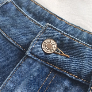 Close-up of blue denim shorts waistband with silver JEANS button and golden stitching