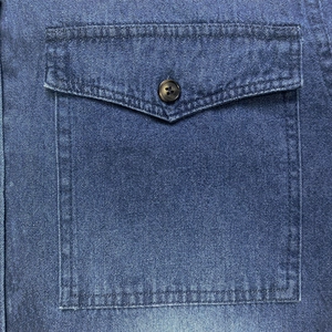 Dettaglio della tasca di una camicia di jeans con bottone marrone scuro e cuciture fini