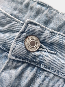 Detail of silver metal button on light blue denim jeans with embossed JEANS text and star