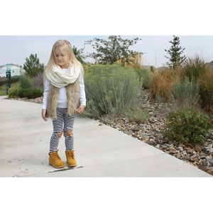 Niña pequeña con chaleco de pelo sintético marrón, bufanda blanca y leggings a rayas