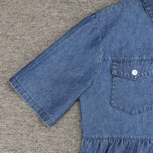 Dettaglio di un abito in denim blu scuro con tasca sul petto, bottone e orlo a pieghe
