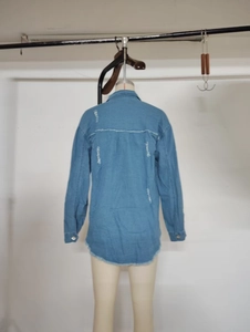Detail view of light blue denim shirt showing distressed holes and frayed raw hem edges