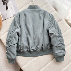 Back view of a sage green bomber jacket with gathered sleeve details on a textured sofa