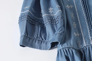 Detailed view of white embroidery and vertical pleats on blue denim dress fabric