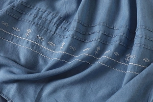 Detailed view of blue denim dress featuring white folk embroidery and pintuck texture