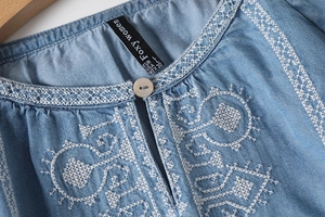 Detail of white embroidery on a light blue denim neckline with brand label and button