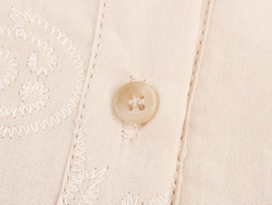 Detail of beige sleeveless dress featuring intricate white embroidery and button closure
