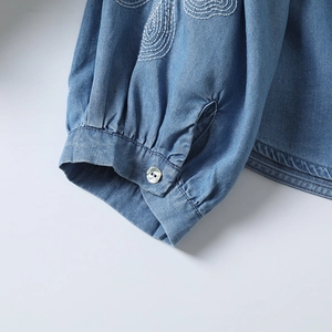 Détail du poignet en denim bleu avec broderie spirale blanche et bouton perlé