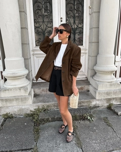 Close-up of a women's brown suede blazer styled with a beige woven handbag and sunglasses