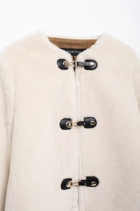 Detail of cream faux fur jacket featuring black leather straps and gold metal buckles