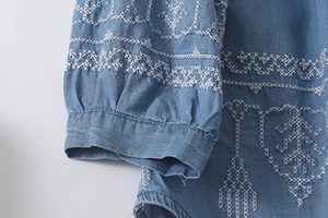 Detail of white cross-stitch embroidery and pleated cuffs on blue denim shirt blouse