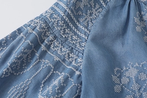 Detailed close-up of white floral embroidery on light blue denim shirt sleeve fabric