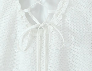 Detail of white lace dress featuring floral embroidery, ruffled neckline, and drawstring bow