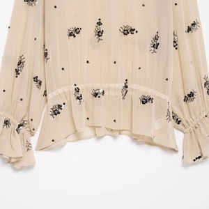 Detail of sheer beige chiffon blouse with black sequin flowers and ruffled hemline