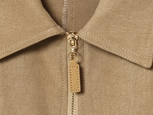 Dettaglio macro della zip dorata con tirante in pelle marrone su giacca twill kaki
