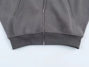 Detail shot of a grey ribbed hoodie hem featuring a metal zipper and side pocket seams