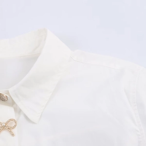 Detail of white poplin shirt featuring pearl metal buttons and a golden rhinestone bow