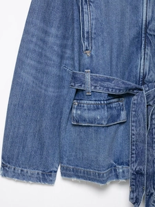 Detail of blue denim jacket showing waist belt, flap pocket with rivets and raw frayed hem