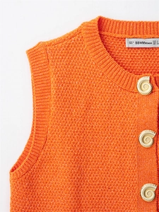 Detail of orange knit vest with textured fabric and golden seashell-shaped buttons