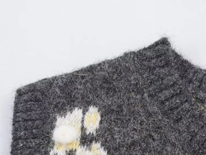 Dettaglio macro di un gilet in maglia grigio scuro con pompon 3D bianchi e fiori