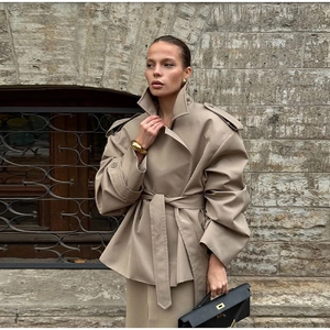 Detail of khaki cropped trench coat with notch lapels, belt, and gold jewelry styling