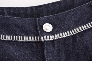 Detailed close-up of dark denim waist with silver metal button and white contrast stitching