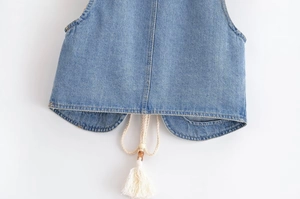 Détail de l'ourlet du gilet en jean bleu clair avec cordon tressé, perles en bois et pompons