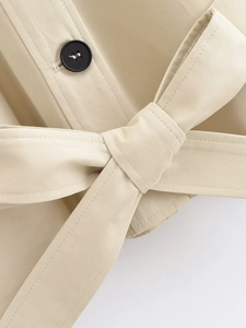 Detail of beige trench coat featuring twill texture, black button, and knotted waist belt