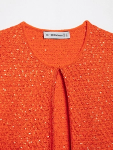 Close-up of bright orange knitted top with gold sequins and BBWMWOMAN brand neck label