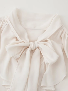 Dettaglio della camicetta in chiffon crema con grande fiocco al collo e spalle plissettate