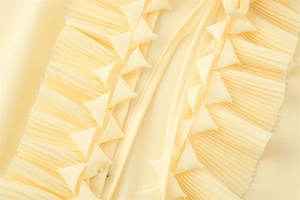 Makro-Detail der Plissee-Rüschen und geometrischen Akzente an einer hellgelben Bluse