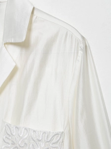 Detail of white satin shirt featuring collar and intricate embroidery on chest pocket