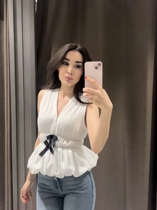 Detail of white V-neck sleeveless peplum top featuring a black bow tie at the waist