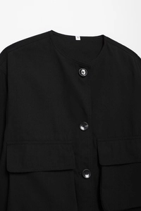 Détail d'une veste noire minimaliste sans col avec texture mate, boutons et poches