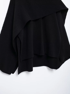 Detail of black knit poncho sweater showing asymmetrical layered drape and side slit hem