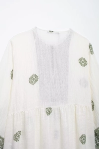 Detailed close-up of white linen fabric with green geometric embroidery and pleated round neck