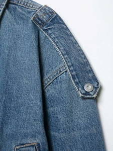 Detail of blue denim jacket showing shoulder strap with metal button and orange stitching