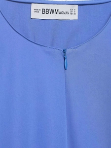 Detail eines hellblauen Hemdausschnitts mit verdecktem Reißverschluss und Markenetikett