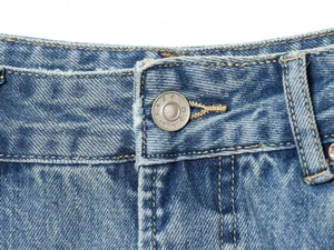 Detailaufnahme eines Silberknopfes und ausgefranster Denimkante an blauen Jeans