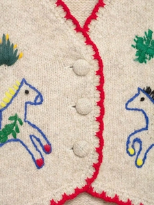 Macro shot of knit vest featuring red scalloped edges and embroidered horse detail