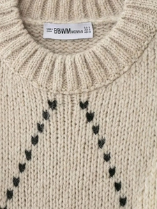 Gros plan du col de pull en maille beige avec motifs de cœurs noirs en losange