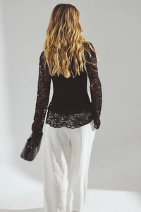 Back view of a model in a sheer black lace top with flared sleeves and white trousers