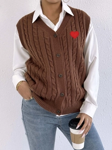 Brown cable knit V-neck vest with red heart patch over a white shirt and blue jeans