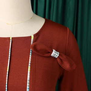 Detail of terracotta dress with square neckline, bow decor, and blue-green plaid trim edges