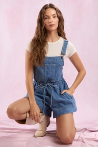 Detail of blue denim overalls with front pocket and drawstring waist on light pink backdrop