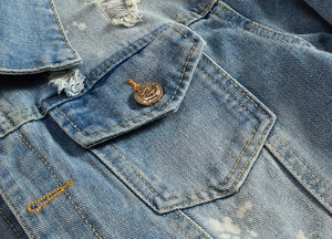 Detail of light blue denim jacket featuring a brass anchor button and distressed pocket edge