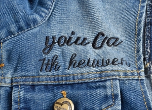 Macro detail of blue washed denim vest with black embroidery and gold fashion button