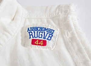 Detail of white denim vest featuring raw fringe edges and colorful brand embroidery
