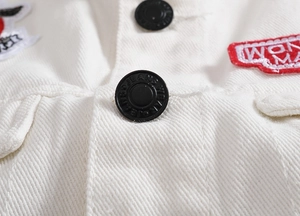 Macro detail of white denim vest featuring black metal buttons and red embroidered patches